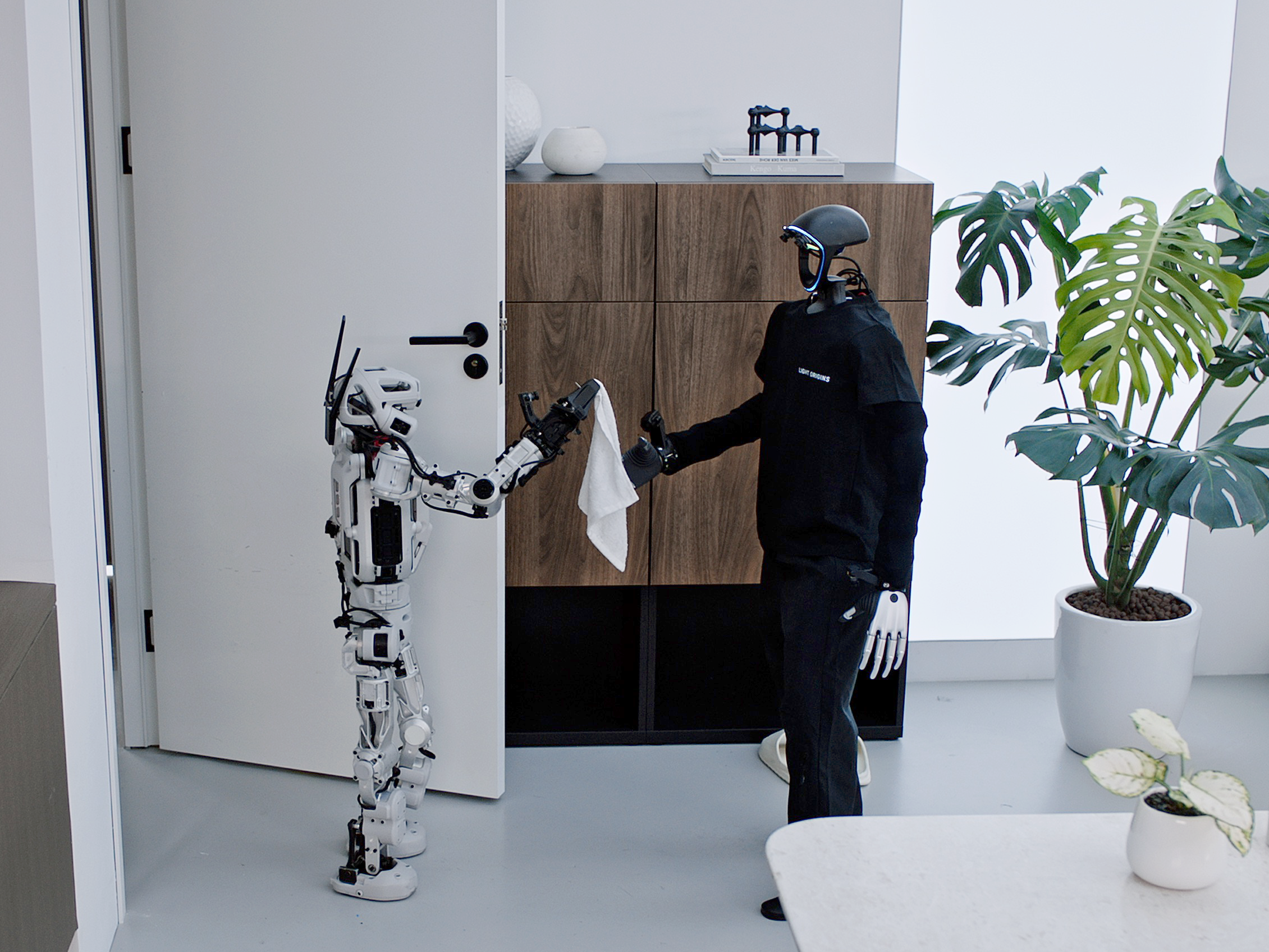 LightBot and another humanoid robot exchanging a white cloth in a furnished room
